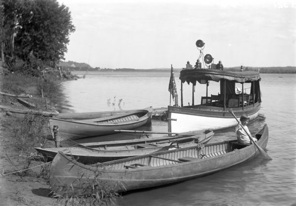 View from shoreline of small launch with two canoes and a rowboat on the shoreline of the Mississippi River. There is a child in the canoe in the foreground.