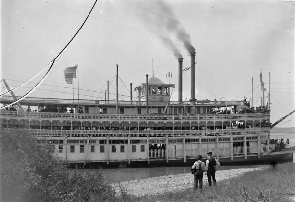 View from shoreline of steamboat. The letters 'J.S.' can be seen at the top of the boat. There are many passengers on board. Two men stand in the foreground on the shore looking at the three-decked sternwheeler.