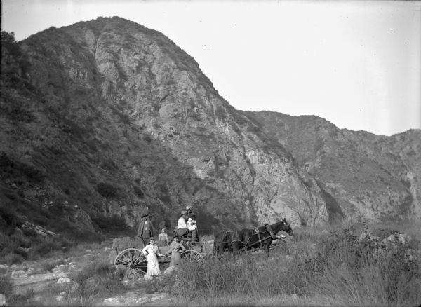 A family of seven, including a baby, posing on and around a wagon with bales of hay in rocky terrain. There are steep hills behind them.