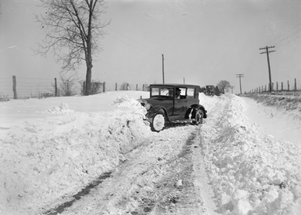 Winter scene with a plowed road and automobile stuck in a snowbank. There is a snowplow in the background.