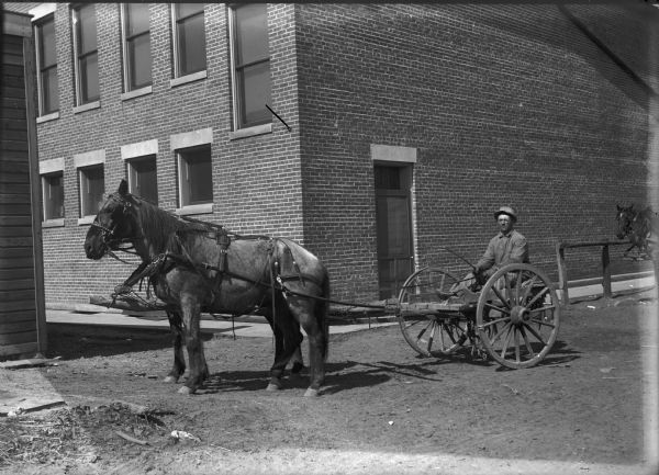 A farmer is squinting against the sun while sitting on a low seat on a two-wheeled cart. Another team of horses are on the right. A large brick building is in the background.