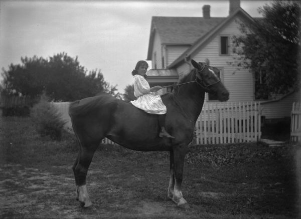 Girl wearing a dress posing on a horse in front of a house.