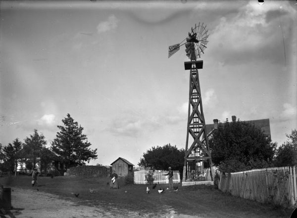 Three people stand in the farm yard. The man on the right is feeding chickens, a woman in the center stands holding an apron. The man on the left is carrying buckets. There is a large building to the left of the woodpile. A fence surrounds the house and workshed. A windmill in the right foreground has writing on one of the blades that says: "Samson The Stove Mfg. Co. Freeport, Ill."