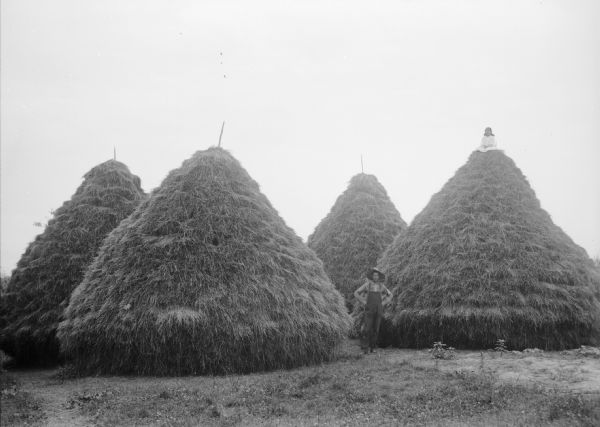 A man is standing among four large haystacks, and a young girl is siting posing on the top of the haystack on the right.