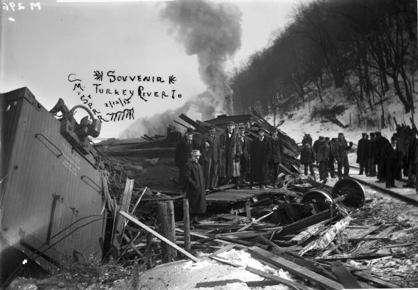 View of group of men standing around the train wreck of the Chicago, Milwaukee, St. Paul and Pacific Railroad near Turkey River, Iowa across the river from Cassville, Wisconsin.