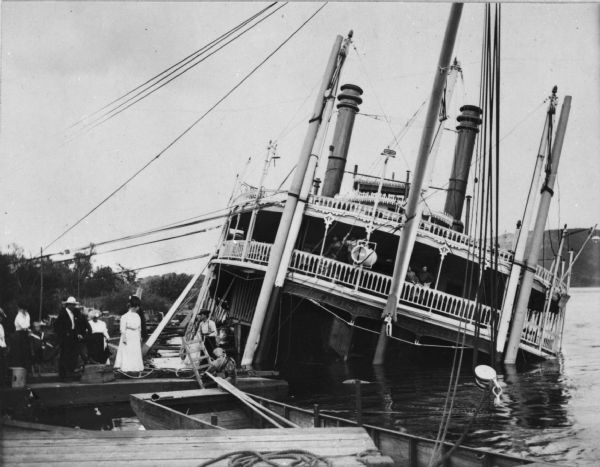 Group of people stand on pier near the Diamond Jo line steamboat <i>Quincy</i>, sunk in 1906. It was raised and renamed the <i>J.S.</i> There are men on the boat, and there is a diver standing on a ladder next to the pier.