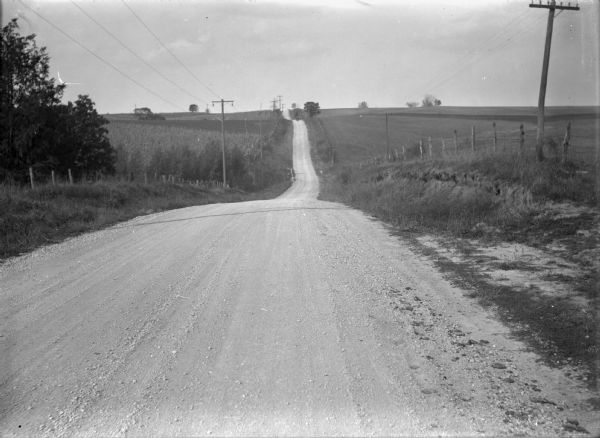 View down hilly unpaved road between two fenced fields. There are telephone poles with wires on either side of the road, and an automible near the top of the next hill.