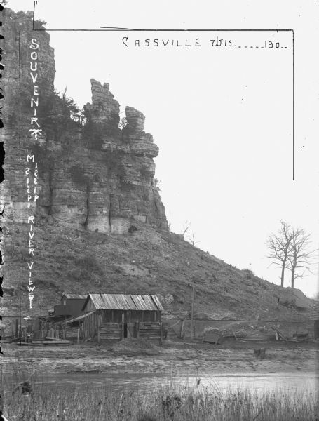 Large rock formation with farm outbuildings, and what appears to be railroad tracks at the base. The Mississippi River is in the foreground. Crop on image depicts how the postcard should be cropped.