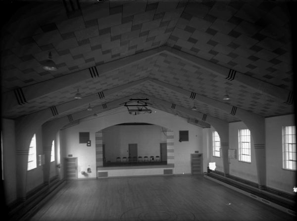Elevated view of gymnasium/theater with vaulted ceiling, large windows along both sides, and a small stage with chairs. There are built-in cupboards or drawers on either side of the stage. A basketball hoop has been retracted up towards the ceiling above the stage.
