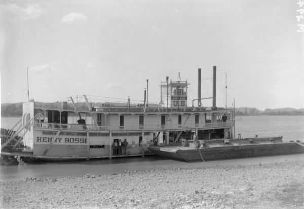 View from shoreline of a group of people standing on the "Henry Bosse" steamboat, a two-decked sternwheeler. The steamboat is attached to a barge along the shoreline.