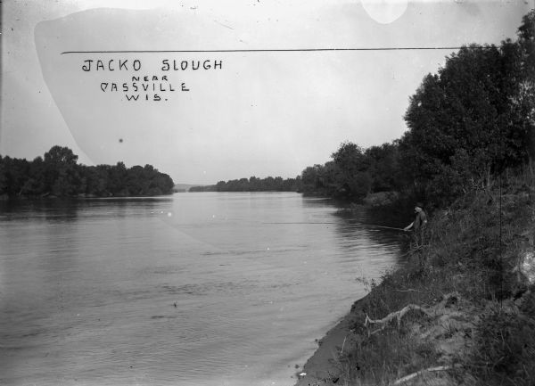 View along shoreline of a man fishing on a bank while smoking a pipe in Jacko Slough.