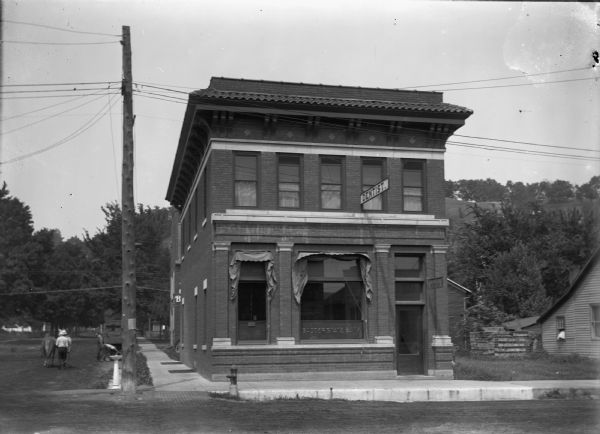 View from street of the Badger State Bank, which is on the first floor. On the second floor is a dentist's office. A telephone or telegraph pole is on the corner, next to what appears to be a water fountain (bubbler). Pedestrians are walking with a horse down the street on the left. In the background on the right is a steep bluff with trees along the top.