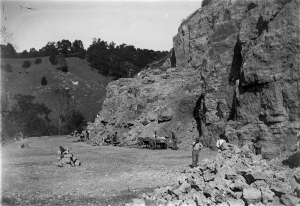 Stone quarry, with men working near a wagon, and at the base of the hill. In the background is a steep hill with trees.