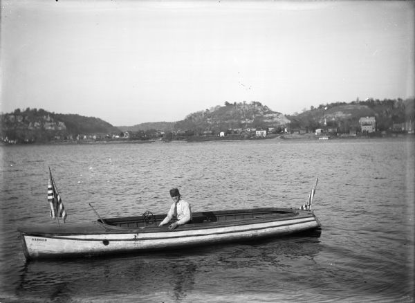 Man sitting in a speed boat named "Badger" on the Mississippi River. There are flags at the stern and bow. In the background is the town of Cassville along the shoreline with bluffs behind.