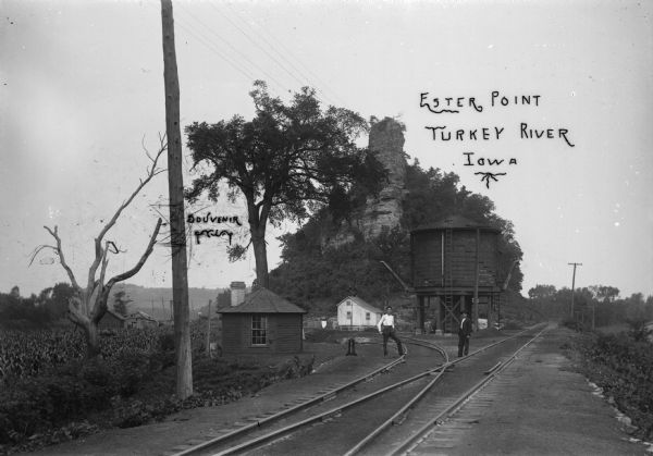 View down railroad tracks, showing the switching station, water tank, and buildings. Two men stand on the railroad tracks. Ester Point is near Cassville, Wisconsin.