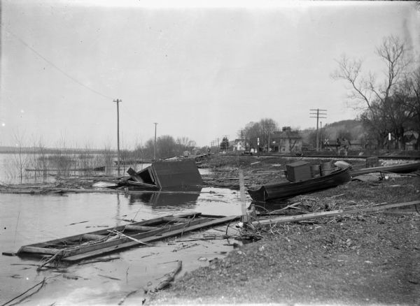 A view along the Mississippi River shoreline after a storm. The water is high and there are damaged boats and debris scattered around. In the background men are standing along the railroad tracks near a handcar.