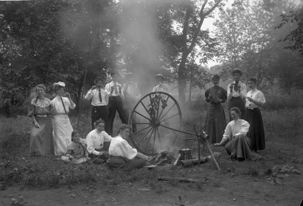 Men and women are having a picnic around a small fire. There is a large wheel for roasting corn. The woman standing farthest to the right is Fannie Kuenster, later Fannie Kuenster Meurer.