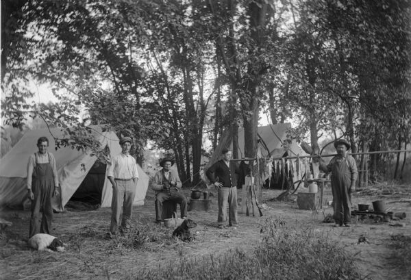 Group portrait of two dogs and five men. In the background are tents among trees. Two of the men are holding a pole strung with their fish catch.