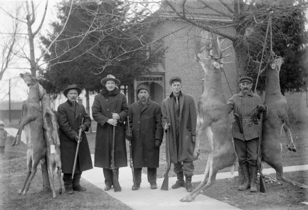 The hunters, holding their guns, are wearing long coats and hats. They are standing on a sidewalk, and the deer are strung up in trees. A small child is standing in the background on the left.