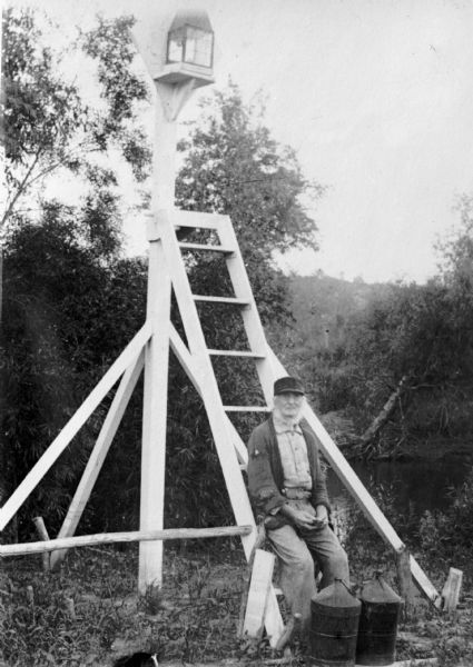 A river navigation light on the Mississippi River.  People were paid to keep them lit so the steam boats could operate at night. Bearded man sitting at base of ladder, which is leaning against a post supporting a lantern. In front of him are what appear to be two containers of lamp oil. There is water and a shoreline in the background.