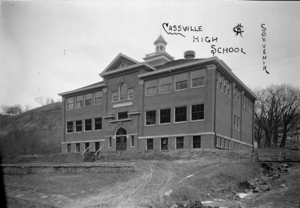 View of facade of high school. The grounds in front are still bare dirt. In the background is a steep hill. A small bridge is over a narrow stream on the right.