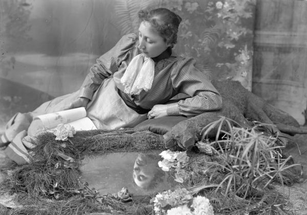 Studio portrait in front of a painted backdrop of a woman posing gazing into a mirror 'pond' while reading a book.