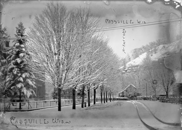 View of snowy street. There are branches down in the road from the trees. In the background is a hill. This is part of Frank Feiker's 'Souvenir' series.