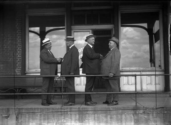Four men are standing at the entrance of what appears to be the Denniston Hotel. The two pairs of men are smiling as they shake hands. Three of them are wearing boater hats. The second man from the left is Charlie Kuenster. Third from the left and back to back with Charlie is his twin brother Henry Kuenster.