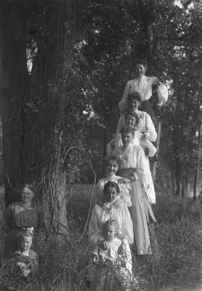 Six young women are posing on a ladder in a woods. There is a young girl in front of them. A woman and a young girl are standing off to the side.