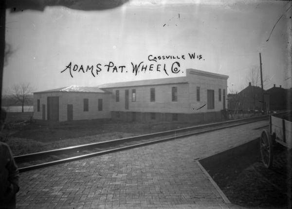 The patent wheel company along the C.B. & Q. Railroad on Front Street. On the far left is a partial view of a person standing close to the camera, and on the right is a cart. In the far background is the Mississippi River and far shoreline.