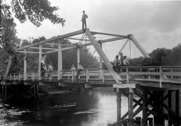 View from shoreline of group of people posing on a bridge across a small river near Cassville, probably in Grant County. The group of people, three women, one child and five men are relaxing comfortably on the bridge. One of the men is fishing from the top of the bridge.