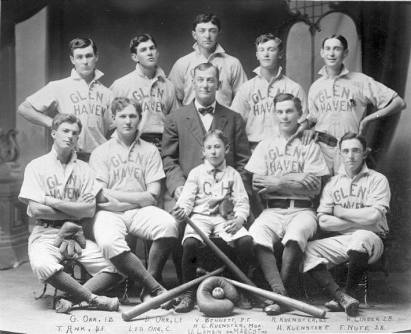 The Glen Haven baseball team in front of a painted backdrop. Names of players: G. Orr, 1st base; T. Ank, center field; D. Orr, left field; Les Orr, Catcher; V. Bennett, right field; H.G. Kuenster, Manager; Eugene Lambin, mascot; R. Kuenster, short stop; H. Kuenster, pitcher; H. Linder, 2nd base; F. Nute, 3rd base.
