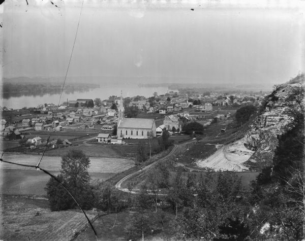 Elevated view over city. A church is in the middle foreground, and the Mississippi River and far shoreline is in the background. On the right side is a dirt road leading to an excavated area below a bluff.