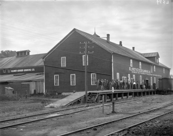 Exterior view of the Klindt-Geiger Canning Company. A group of male and female employees are posing outdoors on the loading dock along the railroad tracks.