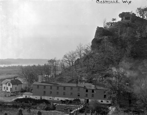 Elevated view of the brewery dwarfed by the overhead bluffs. The brewery was built by William Schmitz. George Scheibl ran the brewery from 1899 until his death in 1904. The brewery operated until 1924, making beer with the trade name "Old Fashion Lager". In 1934, the brewery reopened after the repeal of prohibition and operated until 1938. There is a small house in the lower left of the image. A loaded horse-dawn wagon is ready to go. There are men standing in front of the brewery, and a footbridge in the bottom center of the image. In the far background is the Mississippi River and opposite shoreline.