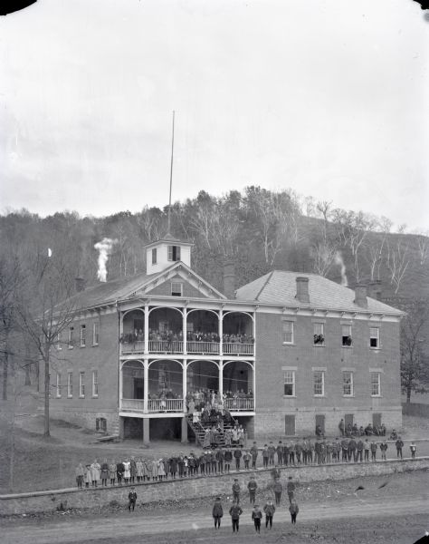 Outdoor portrait of the entire student body posing on the porches and balconies, and on the grounds of the school. A few people are also posing in some of the windows. The school has both a first and second floor porch or balcony. Behind the school is a steep bluff.