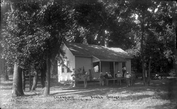 Eight men are posing outside the porch of a cottage which is surrounded by trees. There are blankets and clothes hanging on the clothesline. A work table with a tin washpan is off to the right of the cottage.