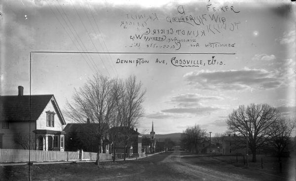 View down Denniston Avenue. On the left side behind a long row of fences along the sidewalk are dwellings and a church. On the right side dwellings are along a footpath. The trees along the avenue are bare of leaves. In the far distance is the Mississippi River and on the far shoreline is a bluff.