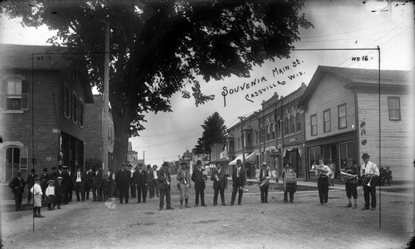One of Frank Feiker's 'Souvenir' photographs. This one is labeled 'no 17' in the left corner and 'no 16' in the right corner. There is a brass band ensemble in the foreground standing in the street. To the left is a group of men and boys. Signs on the storefronts on the left read: "[?]inpell [ware]" and "City Meat Market Kuenster's". Signs on the right read: "Cigar Factory", "Dr DeMers", "Dr. J.B. Curry DDS Dentist" and City Bakery and Restaurant".