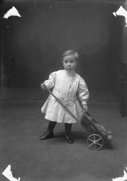 Full-length portrait in front of a painted backdrop of a young child in a dress with puffed sleeves, holding a wagon with a stuffed bear.