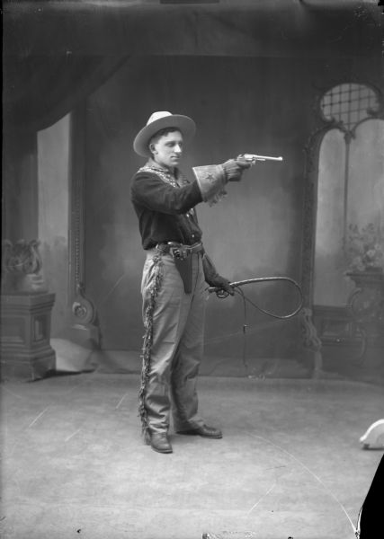 Full-length studio portrait in front of a painted backdrop of a man dressed as a cowboy. He is aiming a gun in one hand and holding a whip in the other. There is another gun in a holster on his belt. He is wearing a hat, pants with fringe along the side, a bandana, and fringed leather gloves decorated with a star.