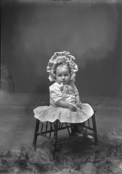 Studio portrait in front of a painted backdrop of a little girl wearing a dress and a frilly hat is posing on a bench. At her feet is a fur rug.