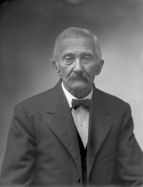 Waist-up studio portrait of an unidentified older man in a suit and bow tie. He has a moustache and white or gray hair.