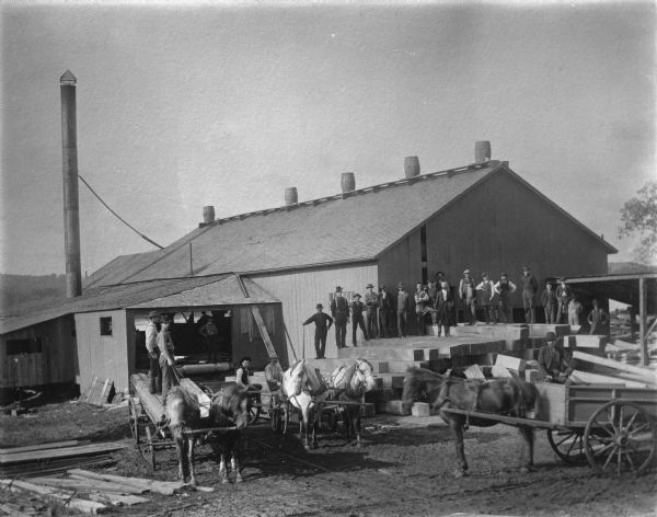 View of men in the Kleinpel lumberyard posing in the midst of work. There are about two dozen men standing on wagons, inside buildings, and on a large stack of wood. There are five barrels on top of the roof of the large building.