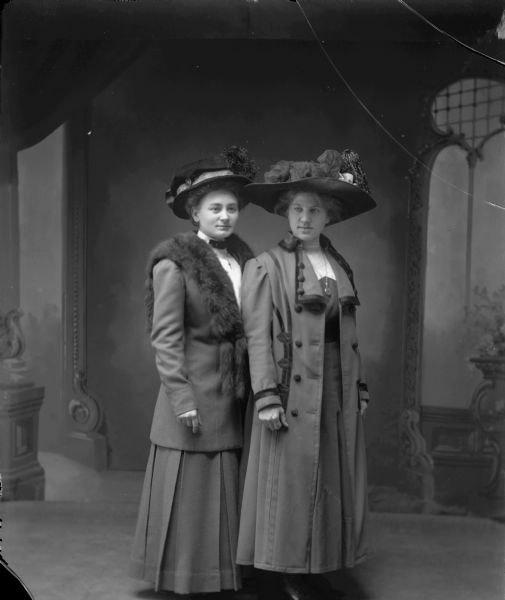 Full-length portrait in front of a painted backdrop of two women wearing fancy hats and tailored suits.
