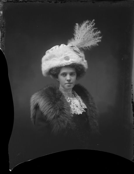 Waist-up studio portrait of a woman in a fur and a lace collar wearing an elaborate light-colored furred and feathered hat.