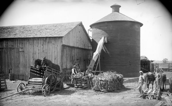 A twenty-five foot high round silo featured on the farm of F.D. Parish, 4.5 miles southeast of Waupaca. The silo is twenty feet in diameter. Two men are getting ready to fill the silo with corn brought by sled. Two horses are standing in a wagon powering what appears to be a belt driven husker and shredder. A conveyor leads to the top of the silo.