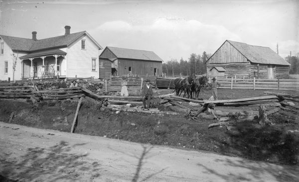 View across road of the log buildings and farm home of August Krueger. Two women and three men stand near the fence along the road. One of the men holds a team of horses.