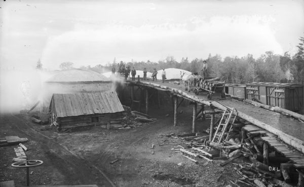 Elevated view of men and horses standing atop the platform used to load wood into charcoal kilns, also known as pits. These kilns convert cord wood into charcoal for furnaces. Smoke surrounds the kiln, and the tops of two other kilns, and railroad cars, are behind the platform.