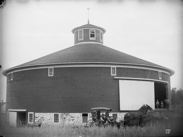 Exterior view of the round, large frame barn of Chas. Tisch. A man in a carriage and a team of horses wearing fly-nets poses in front. On the right a man and two children are standing in the open doorway. The barn measures ninety-two feet in diameter and twenty feet from stone wall to eaves.